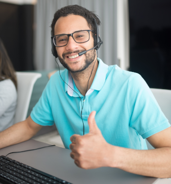 A smiling man in a light blue shirt, sitting at a desk giving a thumbs up gesture.